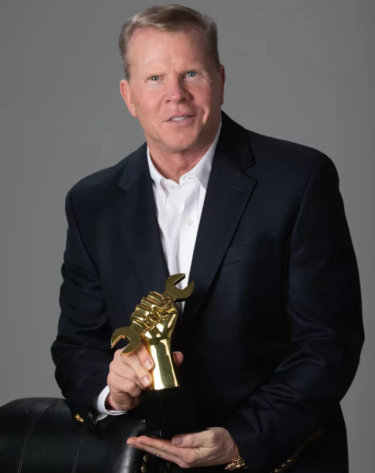 Paul Donahue, the scammer of Advanced Digital Automotive Group, in a black suit, holding the Golden Wrench Award trophy.
